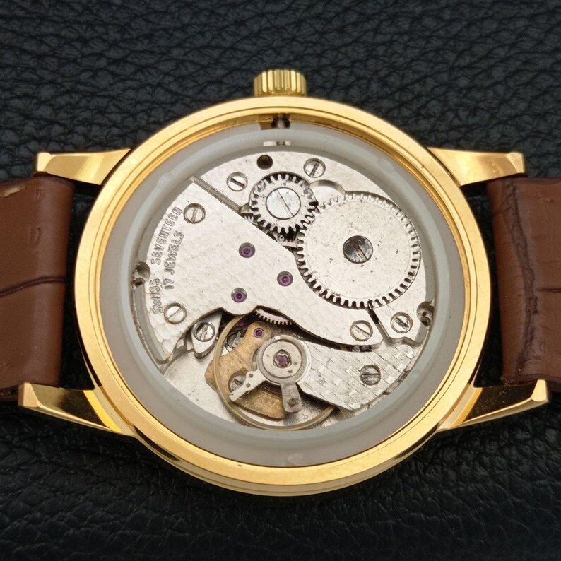 K&ouml;nnte beinhalten: Eine goldfarbene Armbanduhr mit offenem mechanischem Uhrwerk, das durch den Geh&auml;useboden sichtbar ist. Das Werk zeigt silberfarbene Zahnr&auml;der, eine Unruh und die Aufschrift SWISS SEVENTEEN 17 JEWELS. Mit braunem Lederarmband.