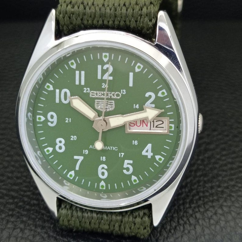 Puede incluir: Un reloj autom&aacute;tico Seiko con esfera verde, caja de acero inoxidable y correa de nailon verde. La esfera muestra el d&iacute;a y la fecha, con agujas e indicadores luminosos. El reloj tiene una est&eacute;tica de estilo militar.