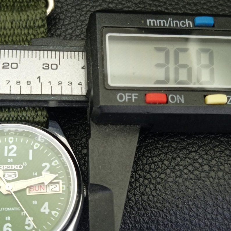 Puede incluir: Primer plano de un reloj Seiko con esfera verde y correa de nailon verde. La esfera muestra la fecha y la hora. Un calibrador digital mide el ancho del reloj en 36,8 mm.