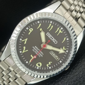 Op de afbeelding: Een zilverkleurig Seiko-horloge met een donkerbruine wijzerplaat en Arabische cijfers. Het horloge heeft een zilveren lunette, een roestvrijstalen band en een dag-datumweergave. Het horloge is gemarkeerd met "660ft = 200m PROFESSIONAL AUTOMATIC".