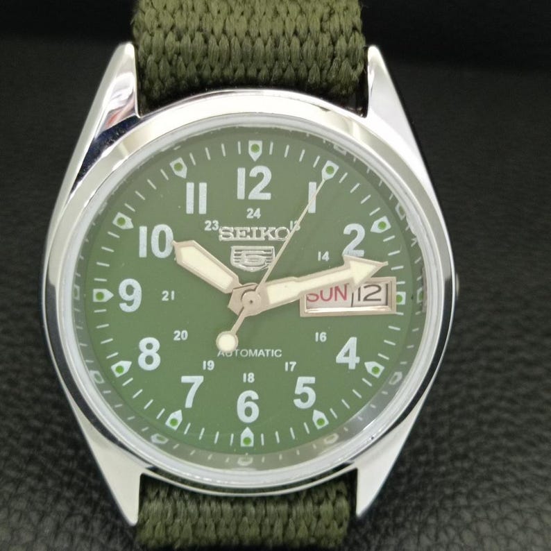 Puede incluir: Primer plano de un reloj autom&aacute;tico Seiko 5 con esfera verde y caja plateada. El reloj tiene una correa de nailon verde. La esfera muestra el d&iacute;a y la fecha.