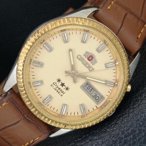 May include: A vintage Orient watch with a cream-coloured face, gold-tone bezel, and a brown leather strap. The watch face displays the day and date, with the text "Crystal 21 Jewels" and the Orient logo.