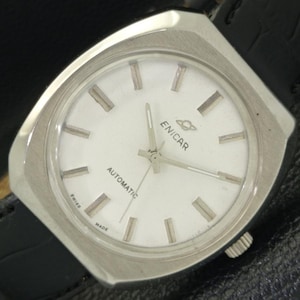 May include: A silver Enicar automatic wristwatch with a white face and black leather strap. The watch face has black hour markers and hands, and the word "Enicar" is printed in black at the bottom of the face.