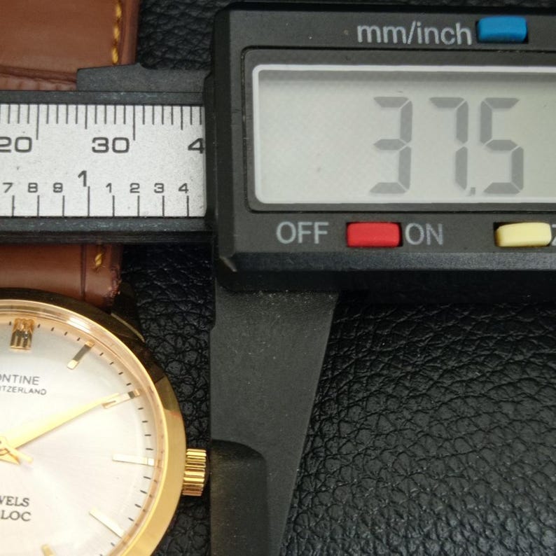 K&ouml;nnte beinhalten: Eine goldfarbene Armbanduhr mit wei&szlig;em Zifferblatt und braunem Lederarmband, neben einem digitalen Messschieber, der einen Wert von 37,5 mm anzeigt. Das Zifferblatt hat goldfarbene Zeiger und Indizes mit dem Text "MONTINE SWITZERLAND" und "JEWELS".