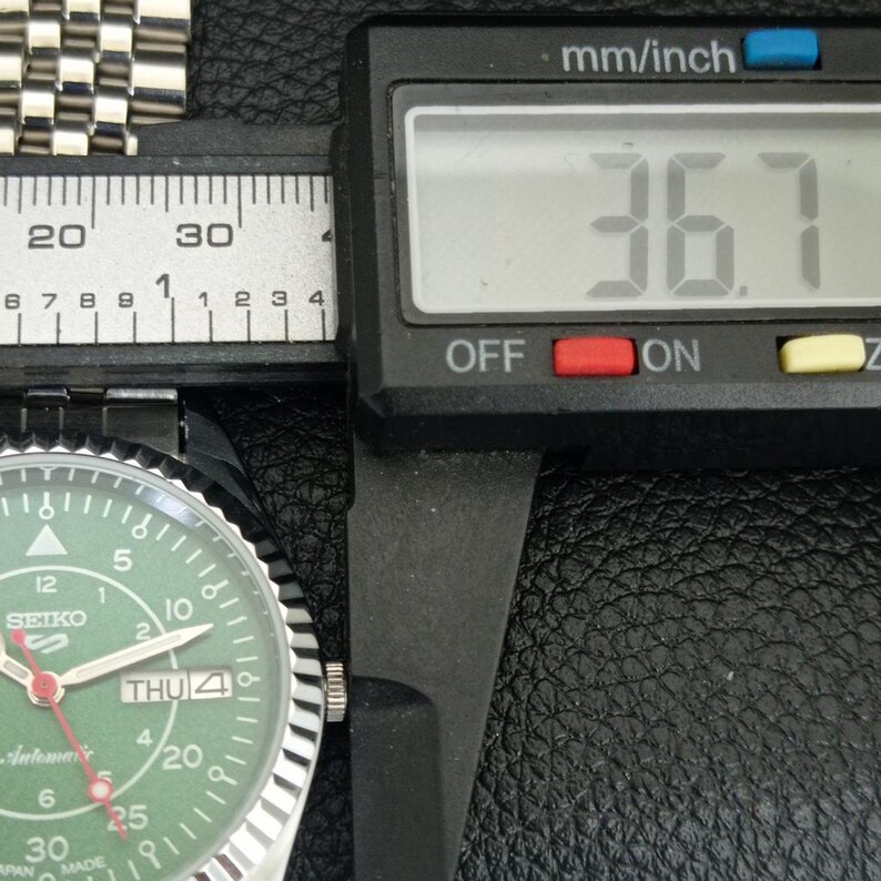 May include: A close-up of a Seiko watch with a green face, silver band, and a digital caliper measuring 36.7 mm. The watch face displays the day and date.