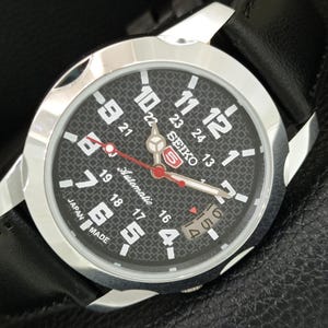 May include: A Seiko 5 automatic wristwatch with a black textured dial, large white Arabic numerals, and a red second hand. The watch features a stainless steel case, a date window at the three o'clock position, and a black leather strap.