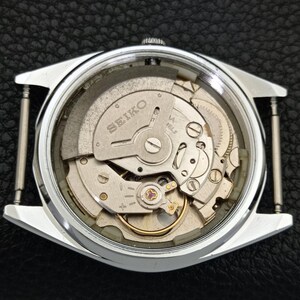 Puede incluir: Vista de cerca de una caja de reloj Seiko plateada, que revela los intrincados mecanismos internos. El movimiento del reloj es visible a trav&eacute;s de una parte trasera transparente, mostrando engranajes, resortes y el logotipo de Seiko. La caja del reloj es rectangular.