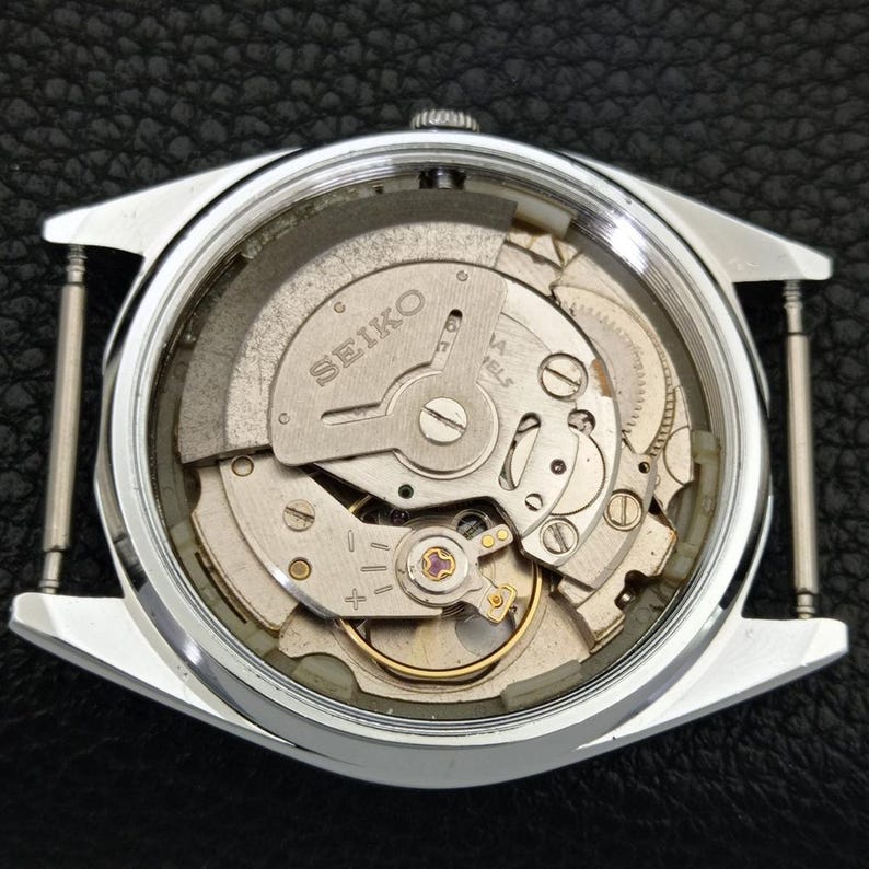 Puede incluir: Vista de cerca de una caja de reloj Seiko plateada, que revela los intrincados mecanismos internos. El movimiento del reloj es visible a trav&eacute;s de una parte trasera transparente, mostrando engranajes, resortes y el logotipo de Seiko. La caja del reloj es rectangular.