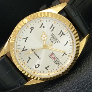 May include: A gold-tone Seiko 5 automatic wristwatch with a fluted bezel, a silver sunburst dial featuring Eastern Arabic numerals, and a black leather strap. The dial displays the Seiko logo, the word Automatic, and a day-date window showing SUN 19.
