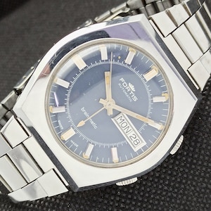 May include: A silver Fortis automatic wristwatch with a blue face, silver hands, and a day and date window. The watch face says "brainmatic" and "MON 28".