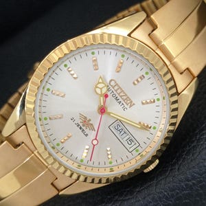 May include: A gold-tone Citizen automatic wristwatch with a fluted bezel and a silver sunray dial. The dial features gold-tone hour markers, a day-date window at the three o'clock position, a red second hand, and the text Citizen Automatic 21 Jewels.