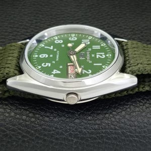 Puede incluir: Un reloj de pulsera plateado con esfera verde y correa de nailon verde. La esfera muestra n&uacute;meros y agujas blancas, con el d&iacute;a y la fecha. El reloj es autom&aacute;tico.