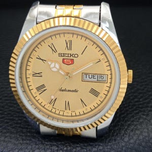 Peut inclure: Une montre automatique Seiko 5 dor&eacute;e avec un bracelet argent&eacute;. Le cadran affiche des chiffres romains, un affichage jour-date et le logo Seiko. Elle poss&egrave;de une lunette cannel&eacute;e et une couronne dor&eacute;e.