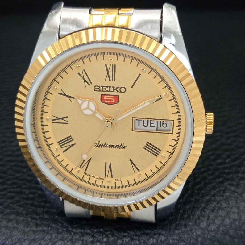 Peut inclure: Une montre automatique Seiko 5 dor&eacute;e avec un bracelet argent&eacute;. Le cadran affiche des chiffres romains, un affichage jour-date et le logo Seiko. Elle poss&egrave;de une lunette cannel&eacute;e et une couronne dor&eacute;e.