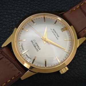 K&ouml;nnte beinhalten: Eine goldfarbene Armbanduhr mit silbernem Zifferblatt im Sonnenschliff, goldenen Zeigern und goldenen Stundenmarkierungen. Das Zifferblatt tr&auml;gt die Aufschrift Montine of Switzerland, 17 Jewels Incabloc und T Swiss Made. Mit braunem Lederarmband.