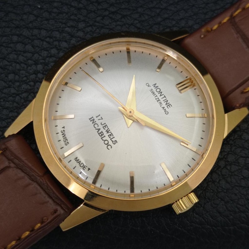 K&ouml;nnte beinhalten: Eine goldfarbene Armbanduhr mit silbernem Zifferblatt im Sonnenschliff, goldenen Zeigern und goldenen Stundenmarkierungen. Das Zifferblatt tr&auml;gt die Aufschrift Montine of Switzerland, 17 Jewels Incabloc und T Swiss Made. Mit braunem Lederarmband.