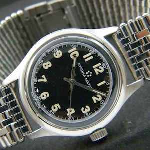 Puede incluir: Un reloj de pulsera con esfera negra, agujas plateadas y números blancos. El reloj tiene una correa plateada y las palabras "ETERNA MATIC" impresas en la esfera.