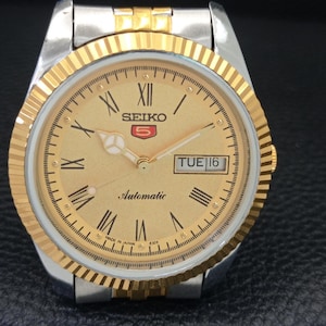 Peut inclure: Une montre automatique Seiko 5 dor&eacute;e avec un bracelet argent&eacute;. Le cadran est dor&eacute; avec des chiffres romains, un affichage jour-date et le mot "Automatic". La montre a une lunette dor&eacute;e cannel&eacute;e.