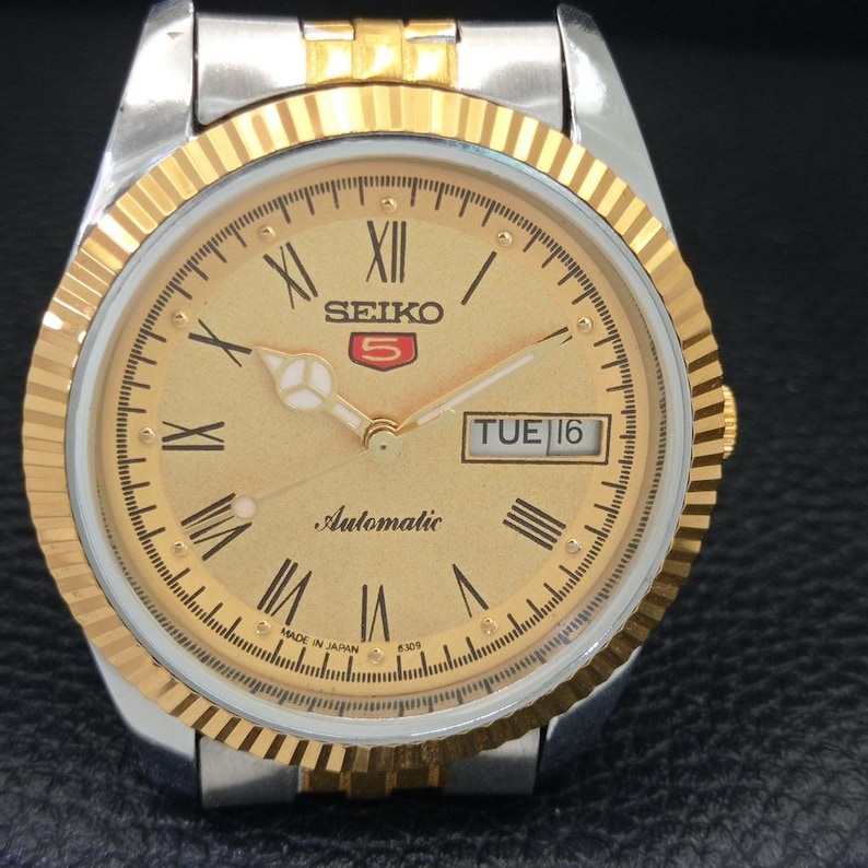 Peut inclure: Une montre automatique Seiko 5 dor&eacute;e avec un bracelet argent&eacute;. Le cadran est dor&eacute; avec des chiffres romains, un affichage jour-date et le mot "Automatic". La montre a une lunette dor&eacute;e cannel&eacute;e.