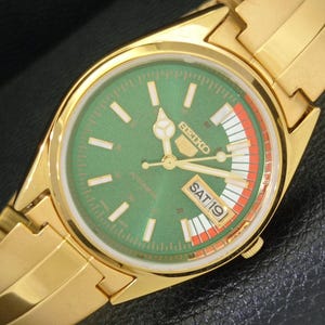 May include: A gold-toned Seiko 5 automatic wristwatch with a green dial. The watch face displays white hour markers, hands, and the day and date. The day is Saturday, the 19th. The watch band is also gold-toned.