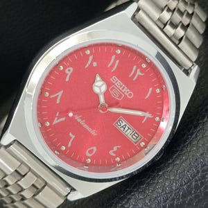 May include: A Seiko 5 automatic wristwatch with a red dial and Arabic numerals. The watch has a silver-tone stainless steel case and band. The day and date are displayed at the 3 o'clock position. The watch is shown on a black surface.