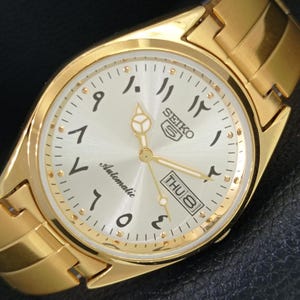 Puede incluir: Reloj de pulsera automático Seiko 5 dorado con esfera plateada y números arábigos. El reloj tiene una pantalla de fecha y una pulsera dorada. La esfera muestra el día como "JUE 8". El diámetro de la caja es de aproximadamente 36 mm.