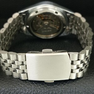 May include: Close-up of a silver wristwatch with a stainless steel band. The watch's back is visible, showcasing the intricate internal mechanisms. The band features a series of small, rectangular links, and the clasp is closed.