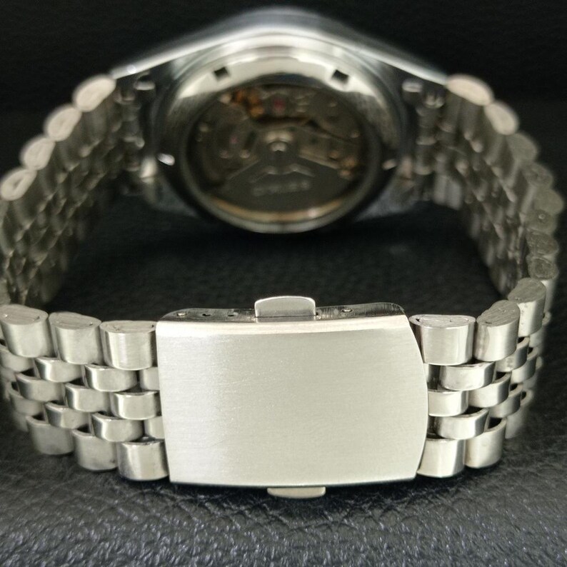 May include: Close-up of a silver wristwatch with a stainless steel band. The watch's back is visible, showcasing the intricate internal mechanisms. The band features a series of small, rectangular links, and the clasp is closed.