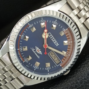 May include: A silver-tone Citizen automatic wristwatch with a blue dial, featuring a fluted bezel, a stainless steel link bracelet, and a day-date window at the three o'clock position. The dial displays the text Citizen, Automatic, and Made in Japan.