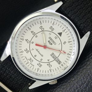 May include: A silver Seiko 5 automatic watch with a white face. The watch has a red second hand, a day-date display, and a black leather strap. The watch face displays the text "SEIKO 5 Automatic" and "Japan Made".
