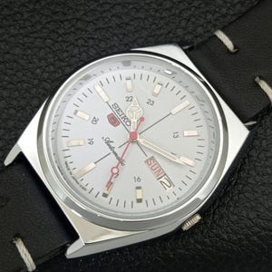 May include: A silver Seiko automatic wristwatch with a white face, black leather band, and day/date display. The watch face features silver hands and markers, with red accents on the second hand and logo. The watch is marked "Made in Japan".
