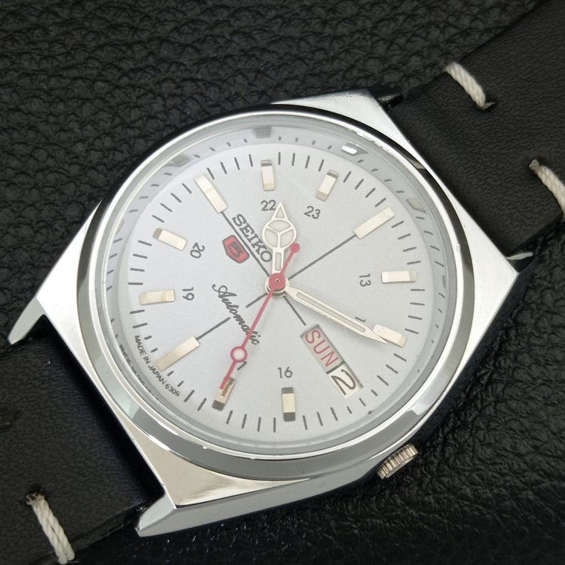 May include: A silver Seiko automatic wristwatch with a white face, black leather band, and day/date display. The watch face features silver hands and markers, with red accents on the second hand and logo. The watch is marked "Made in Japan".