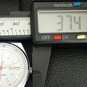 May include: A digital caliper measuring a watch. The display reads 37.4, with the unit of measurement in mm/inch. The watch face is white with black markings and the day is Sunday.