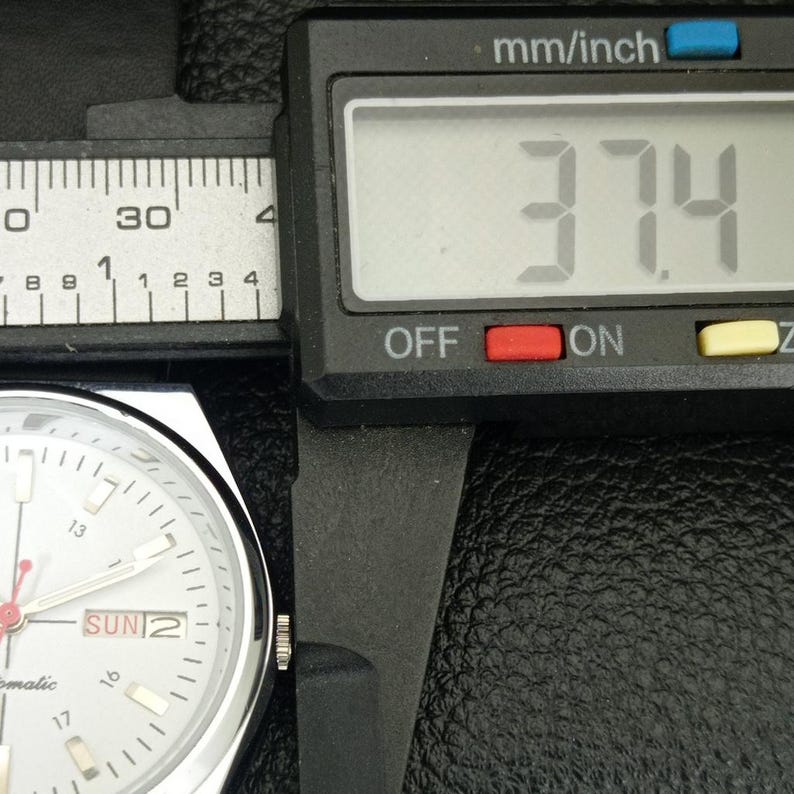 May include: A digital caliper measuring a watch. The display reads 37.4, with the unit of measurement in mm/inch. The watch face is white with black markings and the day is Sunday.