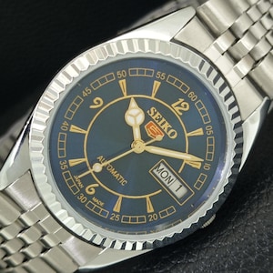 May include: A Seiko automatic wristwatch with a stainless steel band and a dark blue face. The watch face features gold-colored hands, numerals, and markers. The day and date are displayed at the 3 o'clock position. The watch is made in Japan.