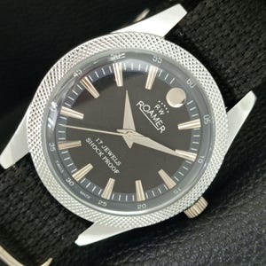 May include: A vintage Roamer wristwatch with a black dial, silver-tone hands, and hour markers. The watch features a textured silver bezel, a black fabric strap, and the text RW ROAMER, 17 JEWELS, SHOCK PROOF, and SWISS MADE on the dial.