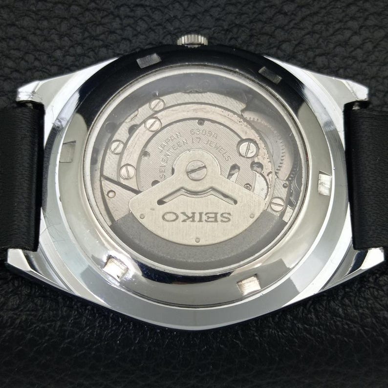May include: Close-up of the back of a silver-toned Seiko watch with a black leather band. The watch's inner workings are visible through a clear glass, showcasing the intricate mechanical movement. The watch face reads "SEIKO" and "JAPAN".