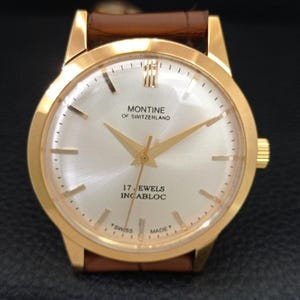 K&ouml;nnte beinhalten: Eine goldfarbene Montine of Switzerland Armbanduhr mit rundem, silbernem Zifferblatt im Sonnenschliff, goldfarbenen Zeigern und Indizes sowie einem braunen Lederarmband. Das Zifferblatt zeigt den Text Montine of Switzerland, 17 Jewels, Incabloc und Swiss Made.