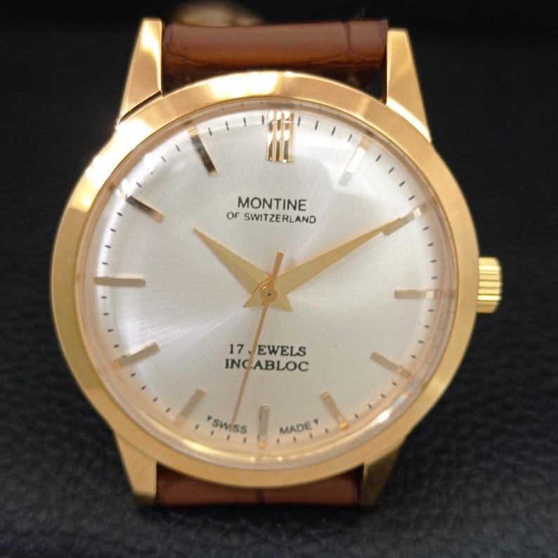 K&ouml;nnte beinhalten: Eine goldfarbene Montine of Switzerland Armbanduhr mit rundem, silbernem Zifferblatt im Sonnenschliff, goldfarbenen Zeigern und Indizes sowie einem braunen Lederarmband. Das Zifferblatt zeigt den Text Montine of Switzerland, 17 Jewels, Incabloc und Swiss Made.