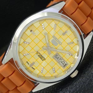 May include: A silver-toned wristwatch with a yellow checkered face and orange rubber strap. The watch face displays the day and date, with the text "AUTOMATIC 17 JEWELS" and "SAT 4". The watch is marked "JAPAN MADE".