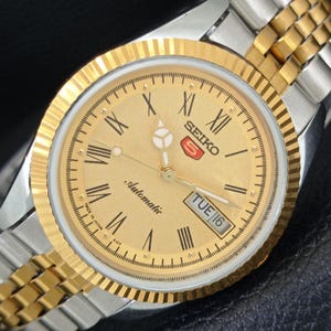 Peut inclure: Montre automatique Seiko 5 dor&eacute;e avec un cadran dor&eacute;, des chiffres romains et un affichage jour-date. La montre a une lunette dor&eacute;e et un bracelet bicolore or et argent. Le cadran indique "Automatic" et "Made in Japan".