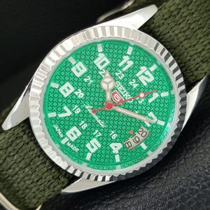 May include: A Seiko 5 automatic wristwatch with a bright green dial featuring a textured geometric pattern, white Arabic numerals, a date window, and a red second hand. The watch has a silver fluted bezel and an olive green fabric strap.