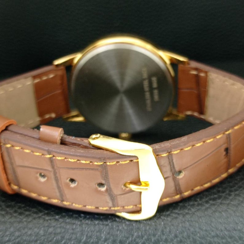 K&ouml;nnte beinhalten: Eine Armbanduhr mit braunem Lederarmband in Krokodilleder-Optik und goldfarbener Schlie&szlig;e. Der Geh&auml;useboden der Uhr ist kreisf&ouml;rmig, aus geb&uuml;rstetem Edelstahl gefertigt und weist eine Gravur auf, die auf den Edelstahlboden hinweist.
