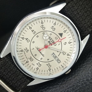 May include: A Seiko 5 Automatic watch with a silver dial, black numerals, and a black fabric strap. The dial features a day-date window at the three o'clock position, a red second hand, and the text Seiko, Automatic, and Made in Japan.