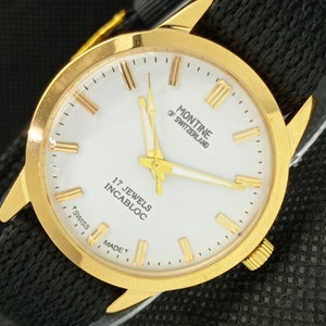 May include: A gold-tone wristwatch with a white face and black band. The watch face has gold-tone hour markers and hands. The watch face also has the text "Montine of Switzerland" and "17 Jewels Incabloc" printed on it. The watch is made in Switzerland.