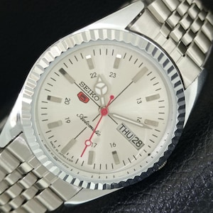 May include: A silver-tone Seiko 5 automatic wristwatch with a fluted bezel and a metallic link bracelet. The off-white dial features silver hour markers, a day-date window at the three o'clock position, and red-tipped hands with the text MADE IN JAPAN 6309.