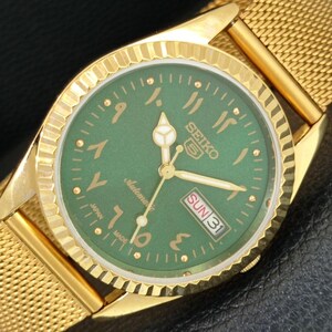 May include: A gold-toned Seiko 5 automatic wristwatch with a green dial. The watch features Arabic numerals, gold hands, and a day-date display. The watchband is a gold mesh style.