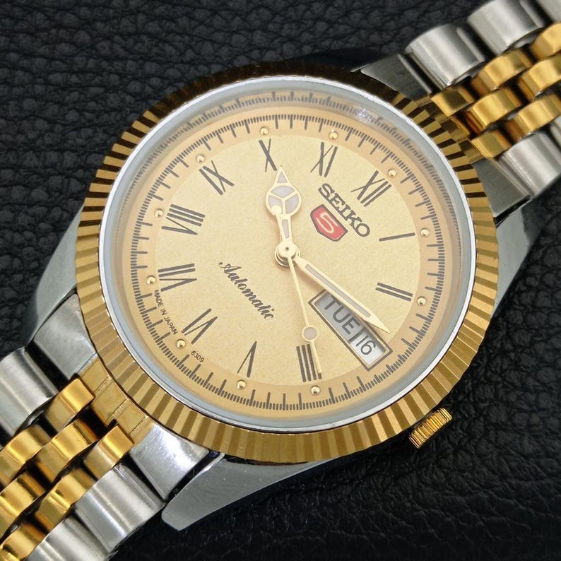 Peut inclure: Montre automatique Seiko 5 dor&eacute;e avec un cadran dor&eacute;, des chiffres romains et un affichage jour-date. La montre pr&eacute;sente une lunette cannel&eacute;e et un bracelet bicolore en acier inoxydable or et argent. Le cadran indique "Automatic" et "TUE 16".