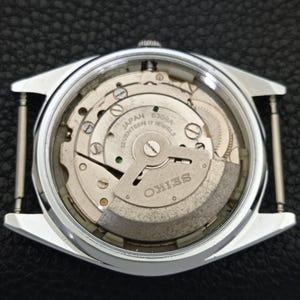 Puede incluir: Una vista de cerca del mecanismo interno de un reloj Seiko. La imagen revela los intrincados engranajes y resortes del reloj, con el nombre de la marca SEIKO claramente visible. La caja del reloj es plateada, rectangular y tiene barras para la correa.