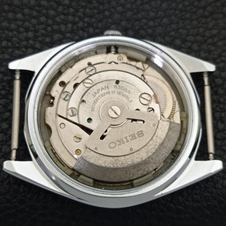 Puede incluir: Una vista de cerca del mecanismo interno de un reloj Seiko. La imagen revela los intrincados engranajes y resortes del reloj, con el nombre de la marca SEIKO claramente visible. La caja del reloj es plateada, rectangular y tiene barras para la correa.